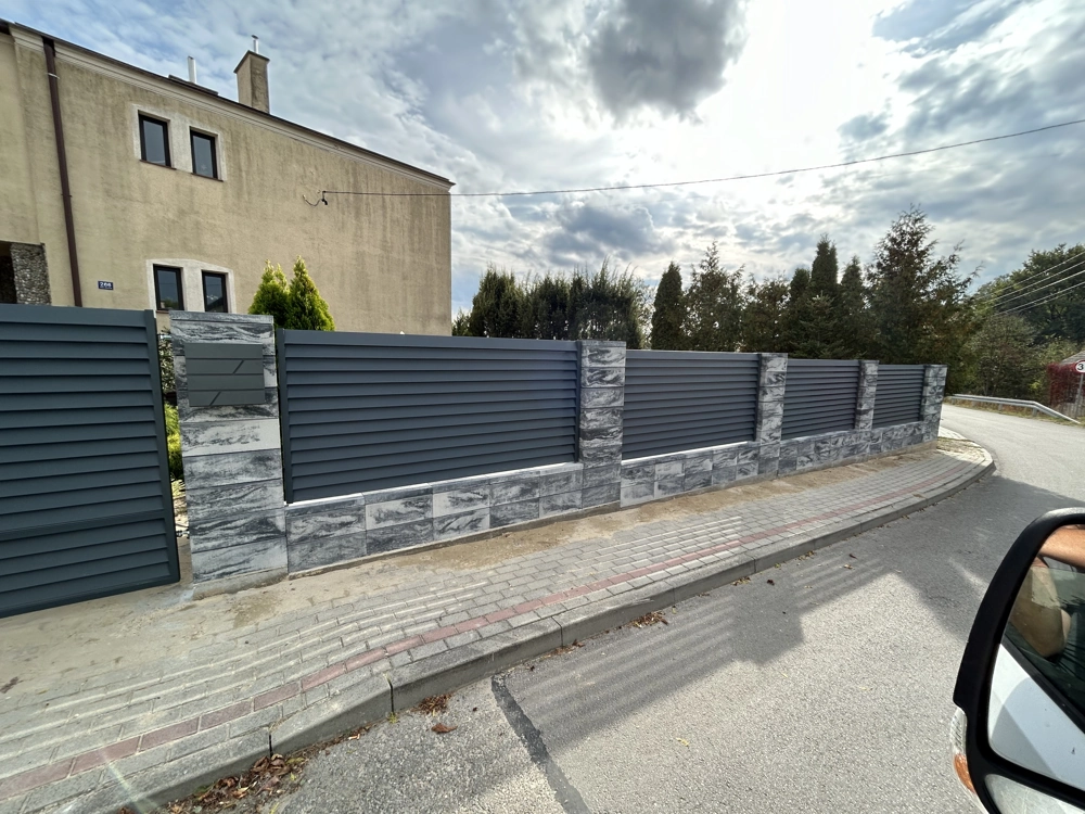 Szare ogrodzenie żaluzjowe z aluminiowych paneli zamontowane na murku z bloczków przy chodniku