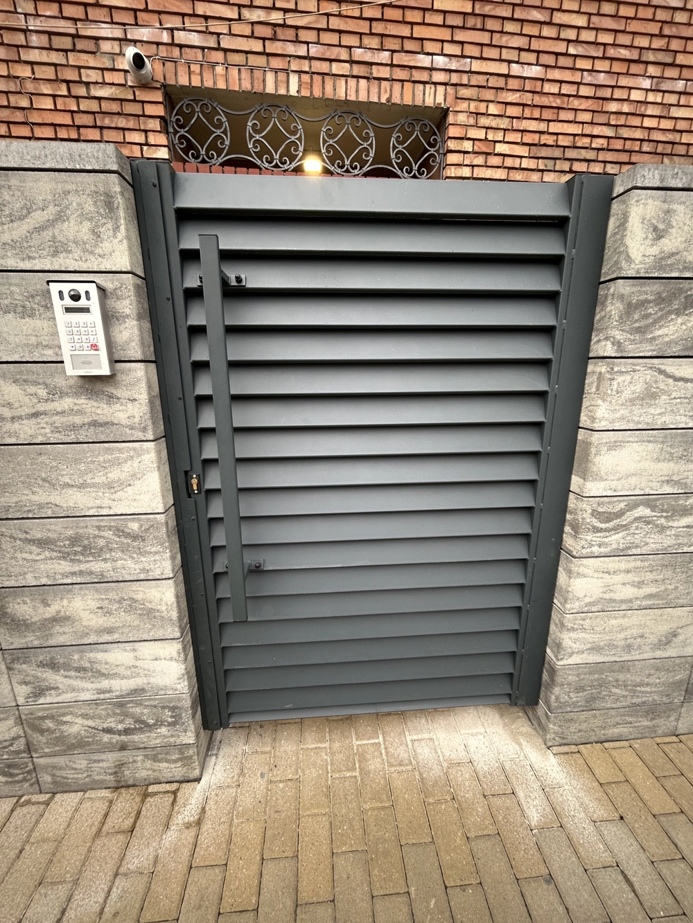 Furtka z poziomych metalowych lameli w stylu ogrodzenia żaluzjowego osadzona w kamiennych słupkach