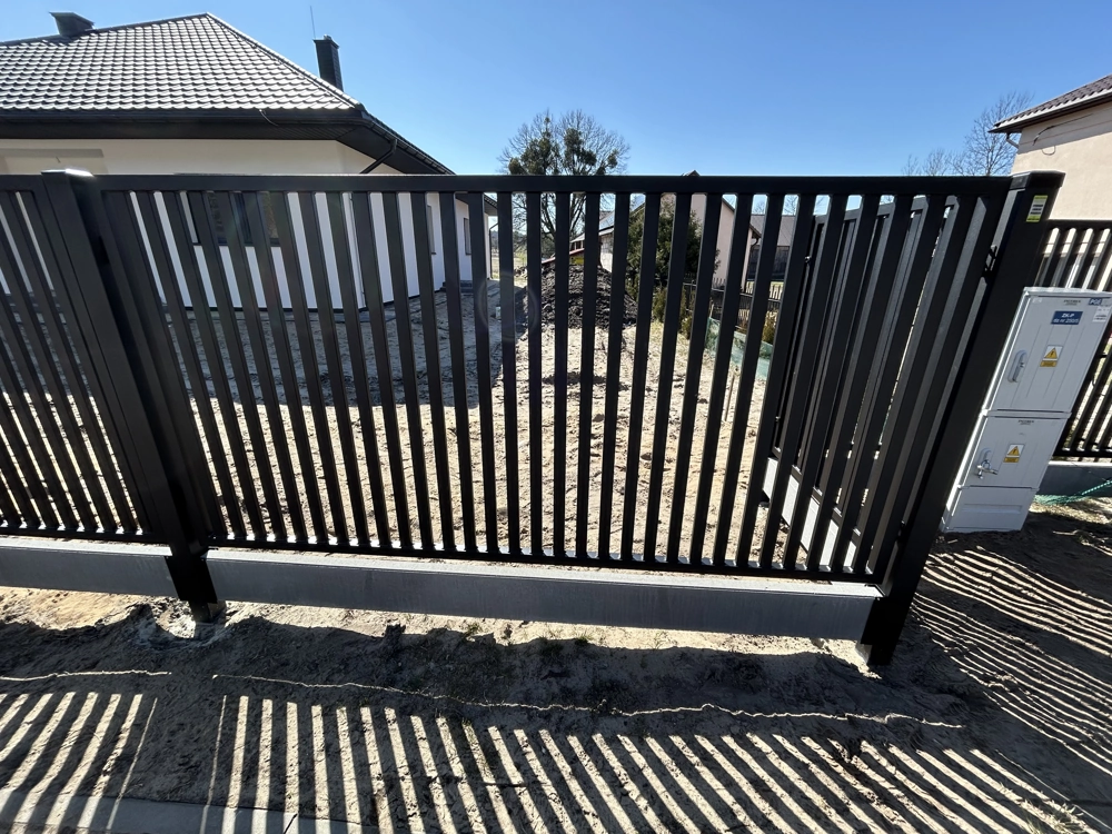 Ogrodzenie palisadowe z czarnych aluminiowych przęseł zamontowane na szarej podmurówce przed domem