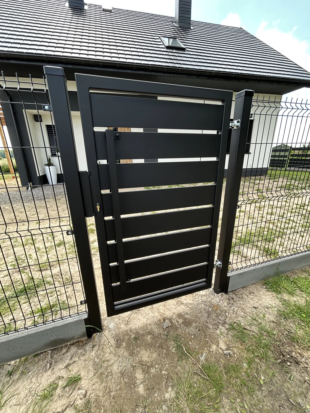 Furtka czarna osadzona w panelowym ogrodzeniu przed nowoczesnym domem