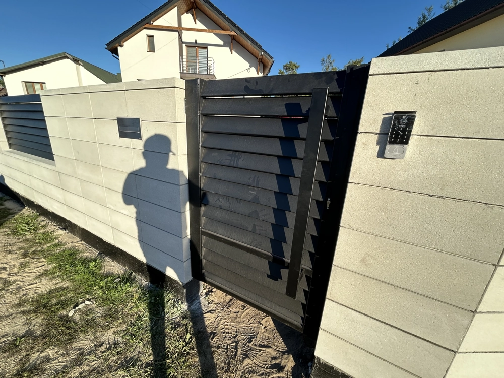Ogrodzenie żaluzjowe: czarna stalowa furtka w betonowym murze z bloczków, z zamontowanym domofonem