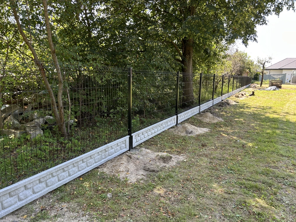 Czarny panel ogrodzeniowy montowany na cokole z szarych bloczków, rozciągający się wzdłuż trawnika i drzew